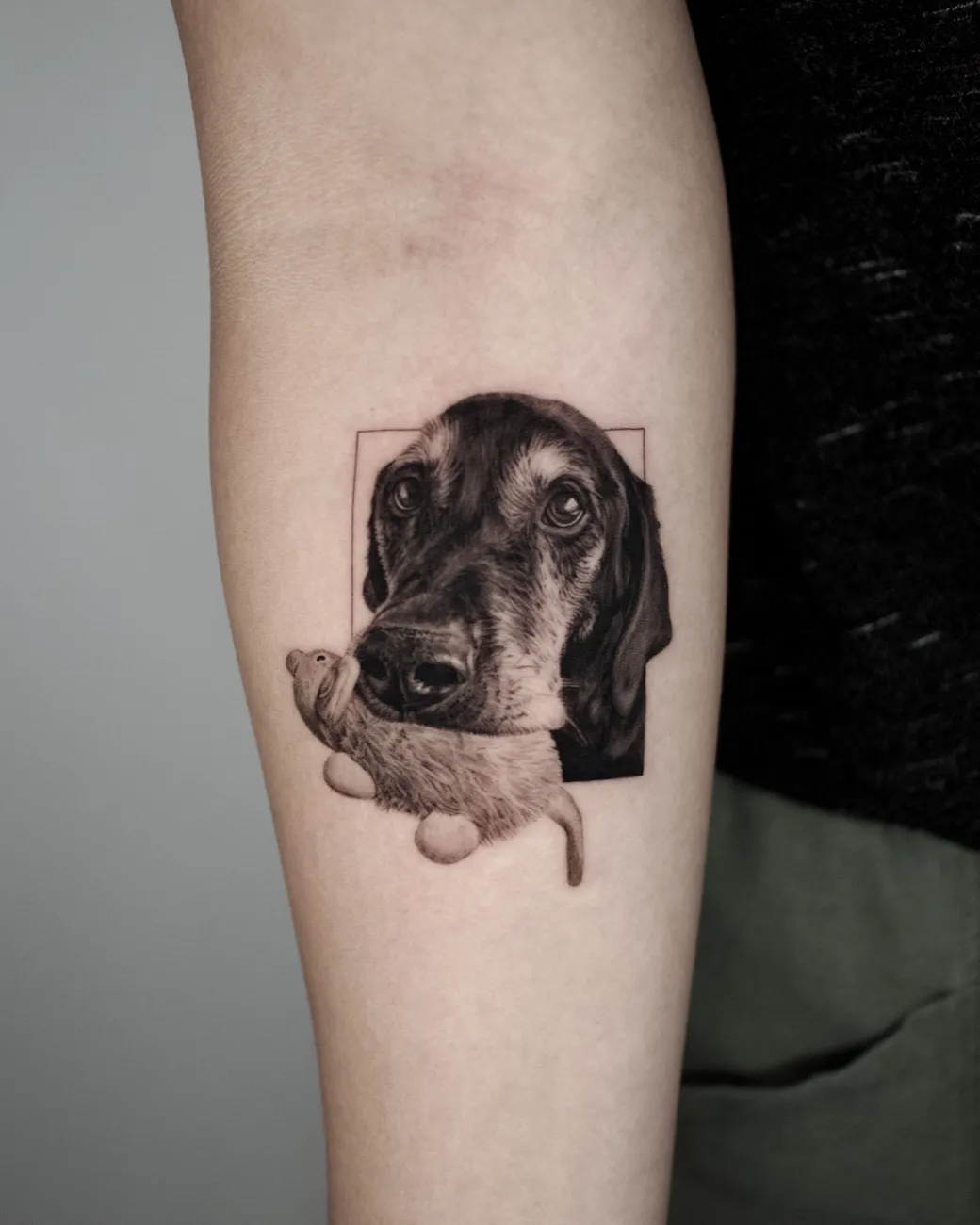 Black and grey realistic tattoo of a dog holding a plush toy